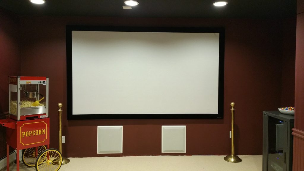 Dedicated home theater by Audio Intellect: a large projection screen over in-wall speakers, dark red walls, a popcorn cart by the door